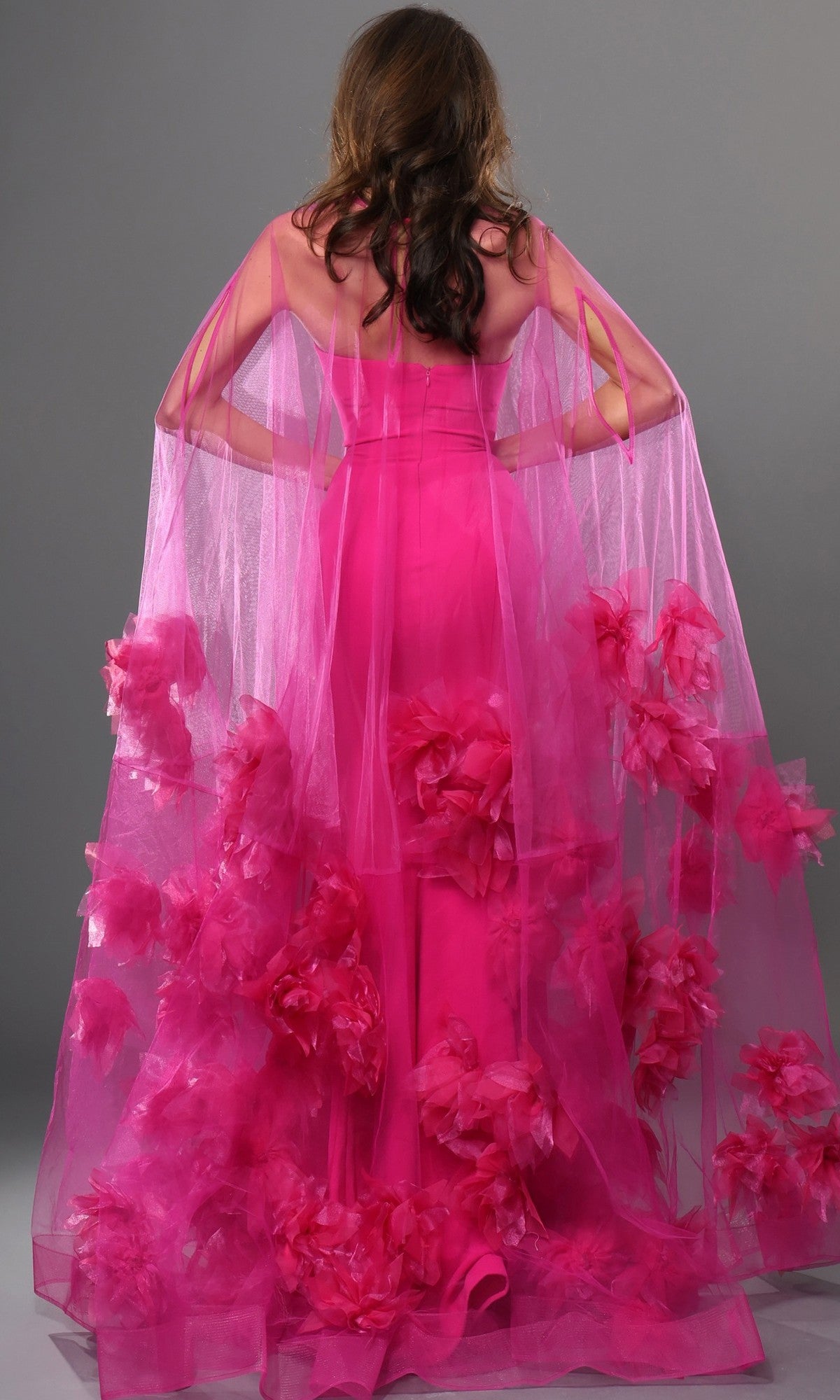 A person is seen from the back wearing the Long Formal Dress: Jovani 40092, a bright pink designer gown with a sheer overlay and large pink floral decorations, standing against a gray background.